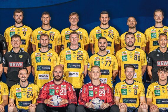 Eine Herren-Handballmannschaft posiert für ein Gruppenfoto. Die Spieler tragen gelbe Trikots, zwei von ihnen haben rote Torwarttrikots an und halten Bälle. Die Trainer und Betreuer in schwarzen Hemden stehen hinter und neben ihnen, alle schauen in die Kamera und lächeln.