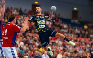 Ein Handballspieler in einem schwarz-gelben Trikot springt in die Luft, um den Ball zu werfen, während Verteidiger in rot-weißen Trikots versuchen, ihn zu blockieren. Das Publikum schaut von der Tribüne im Hintergrund zu.