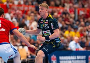 Ein männlicher Handballspieler in einem schwarz-gelben Trikot bereitet sich darauf vor, den Ball zu werfen, während er von einem Gegner in Rot verteidigt wird, mit einer überfüllten Arena im Hintergrund.