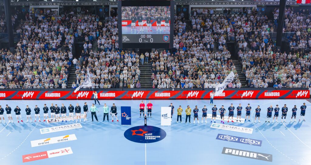 Eine Handballmannschaft stellt sich auf einem blauen Hallenplatz vor einer vollbesetzten Arena mit jubelnden Fans auf, in der Mitte des Platzes stehen Schiedsrichter und Offizielle. Maskottchen halten Fahnen, und im Hintergrund sind eine Anzeigetafel und Werbung zu sehen.