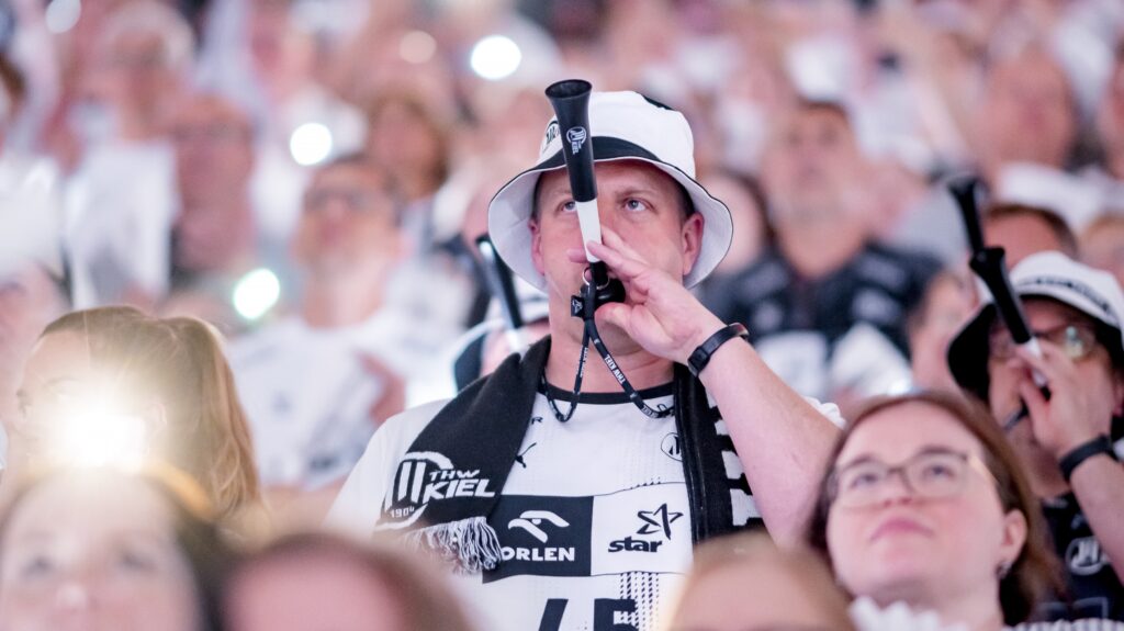 Ein Mann mit weißem Eimerhut und Sporttrikot bläst eine schwarze Vuvuzela inmitten einer Menge von Fans, die alle weiß gekleidet sind, bei einer lebhaften Sportveranstaltung.