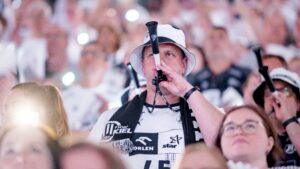 Ein Mann mit weißem Eimerhut und Sporttrikot bläst eine schwarze Vuvuzela inmitten einer Menge von Fans, die alle weiß gekleidet sind, bei einer lebhaften Sportveranstaltung.
