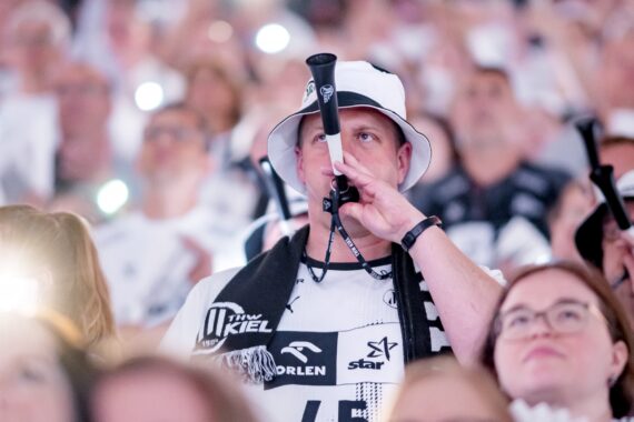 Ein Mann mit weißem Eimerhut und Sporttrikot bläst eine schwarze Vuvuzela inmitten einer Menge von Fans, die alle weiß gekleidet sind, bei einer lebhaften Sportveranstaltung.