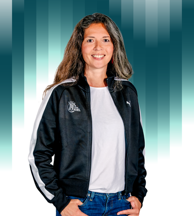 THWKiel-Alisa_Türck Eine lächelnde Frau mit langen braunen Haaren trägt eine schwarze Puma-Jacke und blaue Jeans und steht vor einem Hintergrund mit blaugrünem und weißem Farbverlauf und vertikalen Balken.