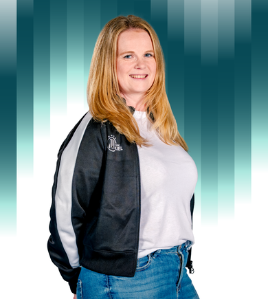 THWKiel-Annika_Lehmann Eine Frau mit langen blonden Haaren, die eine schwarz-weiße Jacke über einem weißen Hemd und eine blaue Jeans trägt, steht lächelnd vor einem gestreiften Hintergrund mit blaugrünem und weißem Farbverlauf.