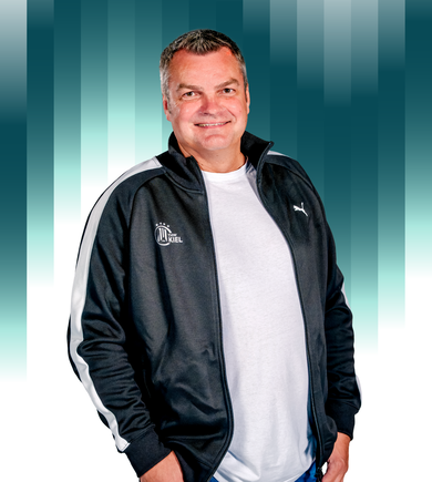Ein Mann mittleren Alters, der eine schwarz-weiße Puma-Sportjacke und ein weißes T-Shirt trägt, steht lächelnd vor einem blaugrün gestreiften Hintergrund.