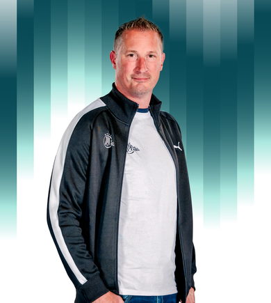 THWKiel-Dirk_Dubbe Ein Mann mit kurzen hellbraunen Haaren, der ein weißes T-Shirt und eine schwarz-weiße Puma-Jacke trägt, steht vor einem geometrischen Hintergrund mit blaugrünem und weißem Farbverlauf.