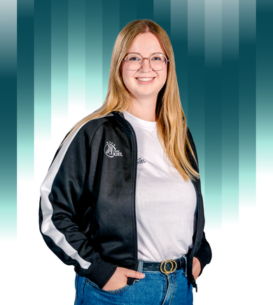 THWKiel-Friederike_Lehmann Eine lächelnde Frau mit langen blonden Haaren und Brille trägt eine schwarz-weiße Sportjacke über einem weißen Hemd und blauen Jeans. Der Hintergrund besteht aus vertikalen blaugrünen und weißen Balken mit Farbverlauf.