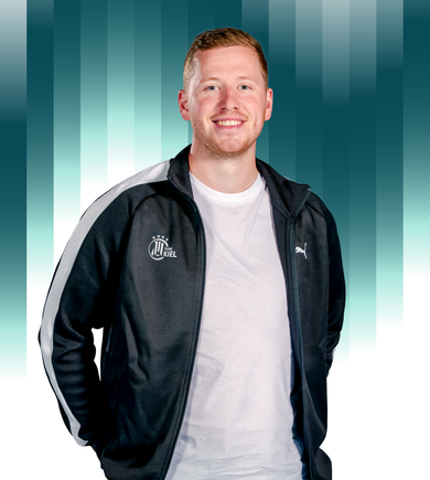 Ein junger Mann in einer schwarz-weißen Puma-Sportjacke mit Kiel-Logo steht lächelnd vor einem blau-weiß gestreiften Hintergrund.