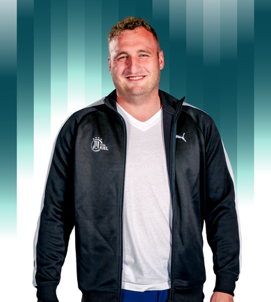 Ein lächelnder Mann in einer schwarzen Puma-Sportjacke über einem weißen T-Shirt steht vor einem Hintergrund mit blaugrünem Farbverlauf und vertikalen Balken. Die Jacke hat ein rundes Logo mit Kiel auf der linken Seite.