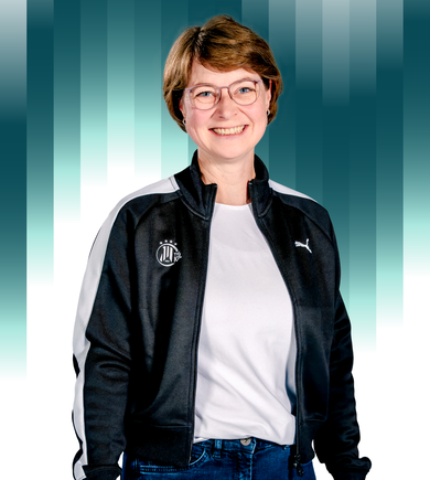 THWKiel-Nina_Andresen Eine lächelnde Frau mit kurzen braunen Haaren und Brille trägt ein weißes Hemd, blaue Jeans und eine schwarze Puma-Jacke mit weißen Streifen und steht vor einem blau-weiß gestreiften Hintergrund.