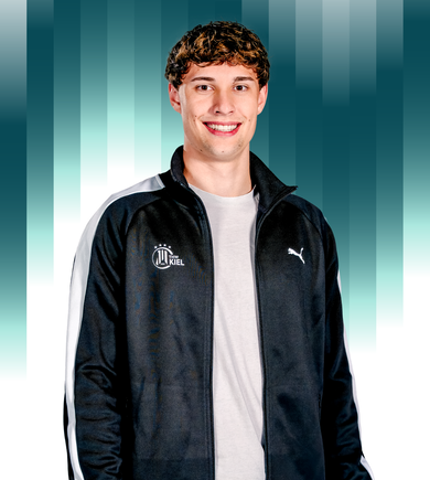 THWKiel-Ole_Westfahl3 Ein junger Mann mit kurzen braunen Haaren, der eine schwarz-weiße Puma-Sportjacke und ein helles Hemd trägt, steht lächelnd vor einem tealfarbenen, senkrecht gestreiften Hintergrund. Auf der Jacke ist ein Logo des THW Kiel zu sehen.