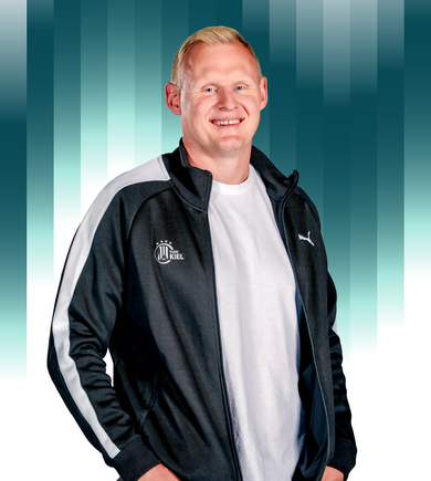 THWKiel-Patrick_Wiencek Ein lächelnder Mann mit kurzen blonden Haaren, der ein weißes T-Shirt und eine schwarz-weiße Sportjacke mit dem THW Kiel-Logo trägt, steht vor einem Hintergrund mit blaugrünem Farbverlauf.