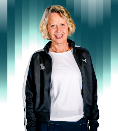 THWKiel-Sabine-Holdurf-Schust Eine ältere Frau mit kurzen blonden Haaren lächelt und trägt eine schwarz-weiße Puma-Sportjacke über einem weißen Hemd. Im Hintergrund sind vertikale Streifen mit Farbverlauf in Grün- und Weißtönen zu sehen.