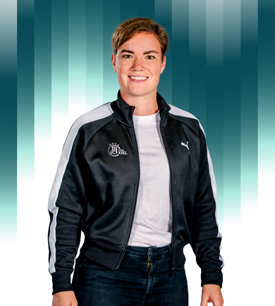 THWKiel-Stefanie_Klaunig Eine Person mit kurzen braunen Haaren, die eine schwarz-weiße Sportjacke mit einem Kiel-Logo und einem Puma-Emblem trägt, steht lächelnd vor einem Hintergrund mit blaugrünem und weißem Farbverlauf.