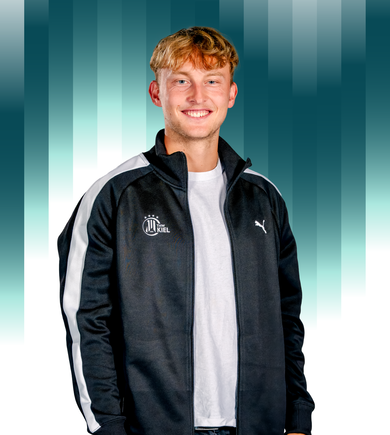 Ein junger Mann mit blondem Haar, der eine schwarz-weiße Sportjacke mit Puma- und Kiel-Logos und ein weißes Hemd trägt, steht lächelnd vor einem blauen Hintergrund mit vertikalen Streifen.