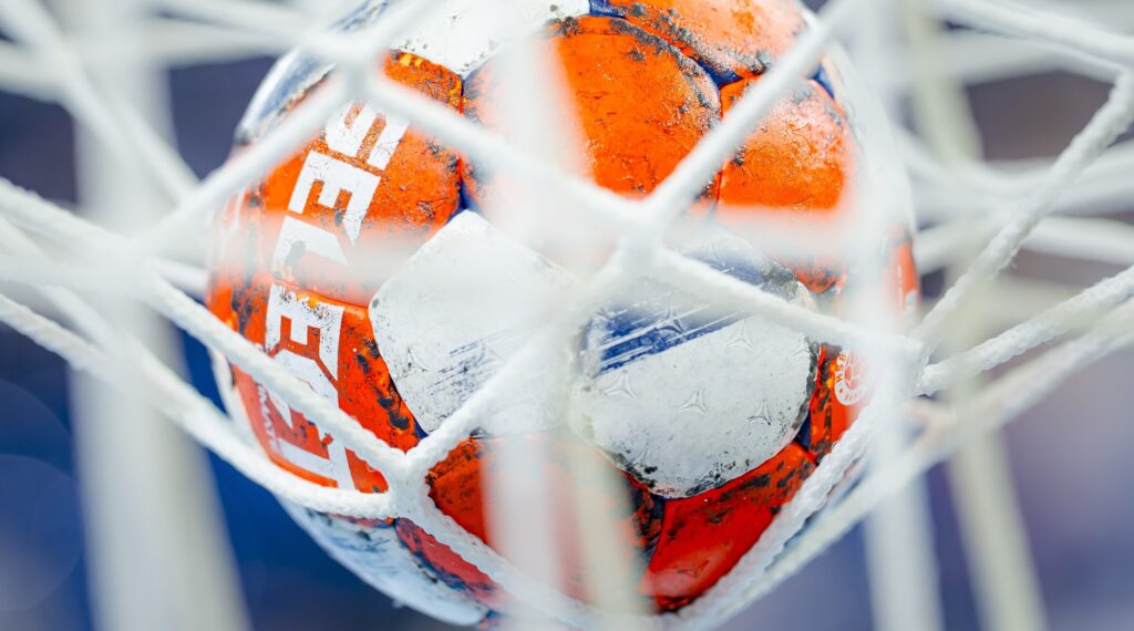 HANDBALL 2024/25: THW Kiel - Limoges Handball Eine Nahaufnahme eines orange-weiß-blauen Handballs, der in einem weißen Netz gefangen wurde. Der Hintergrund ist unscharf, um die Details des Balls und des Netzes hervorzuheben - so wird die Spannung der THW Kiel EHF European League Saison 2025/2026 eingefangen.