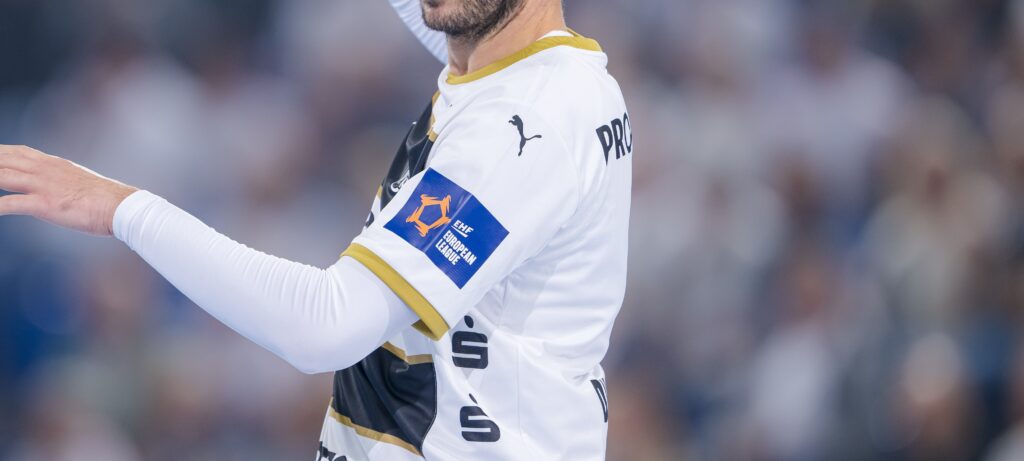 HANDBALL 2024/25: THW Kiel - Bathco BM Torrelavega Ein Handballspieler im weißen Trikot mit goldenen Verzierungen des THW Kiel hebt während eines Qualifikationsspiels der Europa-League-Saison 2025/2026 gegen den BSV Bern den Arm; der Fokus liegt auf seinem Oberkörper und dem Sponsorenaufnäher, während im Hintergrund eine unscharfe Menschenmenge zu sehen ist.