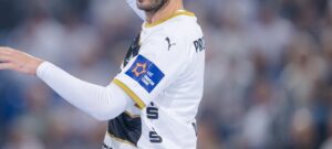 Ein Handballspieler im weißen Trikot mit goldenen Verzierungen des THW Kiel hebt während eines Qualifikationsspiels der Europa-League-Saison 2025/2026 gegen den BSV Bern den Arm; der Fokus liegt auf seinem Oberkörper und dem Sponsorenaufnäher, während im Hintergrund eine unscharfe Menschenmenge zu sehen ist.