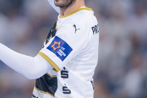 Ein Handballspieler im weißen Trikot mit goldenen Verzierungen des THW Kiel hebt während eines Qualifikationsspiels der Europa-League-Saison 2025/2026 gegen den BSV Bern den Arm; der Fokus liegt auf seinem Oberkörper und dem Sponsorenaufnäher, während im Hintergrund eine unscharfe Menschenmenge zu sehen ist.