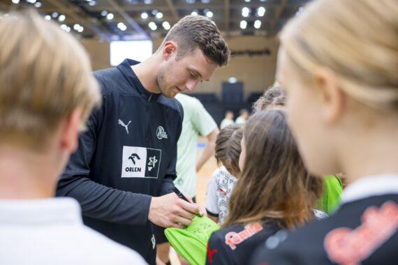 Ein männlicher Sportler in einem schwarzen Sporttrikot gibt einer Gruppe von Kindern in einer Sporthalle Autogramme. Die Kinder stehen dicht bei ihm und halten ihm ihre Trikots zum Unterschreiben hin. Der Hintergrund zeigt eine Menschenmenge und helle Scheinwerfer.