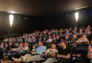Ein überfüllter Kinosaal mit Menschen unterschiedlichen Alters, die in bequemen Sitzen sitzen, Snacks essen und Getränke trinken, alle mit Blick auf die Leinwand und aufmerksam. Die Atmosphäre ist lebhaft und engagiert.