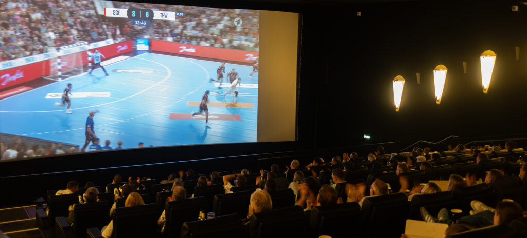 251013_Partner-Viewing_Cinemaxx_Titel Eine große Gruppe von Menschen sitzt in einem dunklen Kino und sieht sich ein Handballspiel an, das live auf die große Leinwand projiziert wird. Das Spiel läuft, und die Anzeigetafel ist sichtbar. Warme Wandleuchten erhellen den Saal.