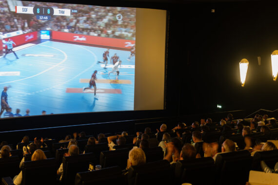 Eine große Gruppe von Menschen sitzt in einem dunklen Kino und sieht sich ein Handballspiel an, das live auf die große Leinwand projiziert wird. Das Spiel läuft, und die Anzeigetafel ist sichtbar. Warme Wandleuchten erhellen den Saal.