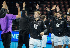 Mehrere männliche Athleten in Sportuniformen klatschen sich auf einem Hallenplatz gegenseitig ab. Ein Spieler, der die Nummer 15 trägt, steht in der Mitte, und im Hintergrund sind unscharfe Zuschauer und leere Plätze zu sehen.