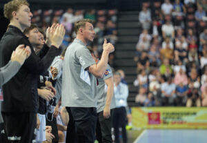 Ein Trainer in einem grauen Polohemd steht am Rande einer Hallensportveranstaltung und zeigt den Daumen nach oben, während Spieler und Zuschauer das Geschehen beobachten. Das Publikum sitzt in Sitzreihen im Hintergrund.