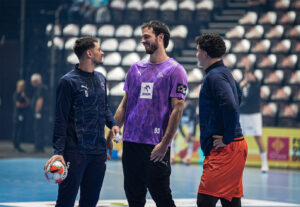 Drei männliche Handballspieler stehen auf einem Hallenplatz, unterhalten sich und lächeln. Einer hält einen Handball in der Hand, ein anderer trägt ein lila Trikot, und die Tribünen hinter ihnen sind fast leer.