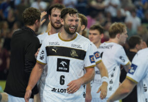 Ein lächelnder Handballspieler in einem weißen Trikot mit der Nummer 6 feiert mit seinen Mannschaftskameraden auf dem Spielfeld, umgeben von anderen Spielern und unscharfen Zuschauern im Hintergrund.