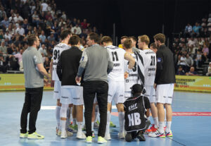 Eine Herren-Handballmannschaft in weißen und schwarzen Trikots kauert auf einem Hallenplatz zusammen, während ein Trainer und ein Betreuer in der Nähe stehen. Ein Fotograf kniet davor, und eine Menschenmenge schaut von der Tribüne im Hintergrund zu.