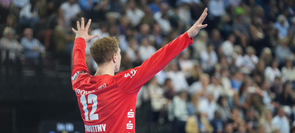 251014_Montpellier_THW_Titel_Nowottny Ein Handballspieler, der ein rotes Trikot mit der Nummer 12 trägt, steht mit erhobenen Armen vor einer verschwommenen Menge in einer Arena.