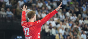 Ein Handballspieler, der ein rotes Trikot mit der Nummer 12 trägt, steht mit erhobenen Armen vor einer verschwommenen Menge in einer Arena.