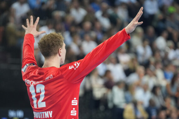 Ein Handballspieler, der ein rotes Trikot mit der Nummer 12 trägt, steht mit erhobenen Armen vor einer verschwommenen Menge in einer Arena.