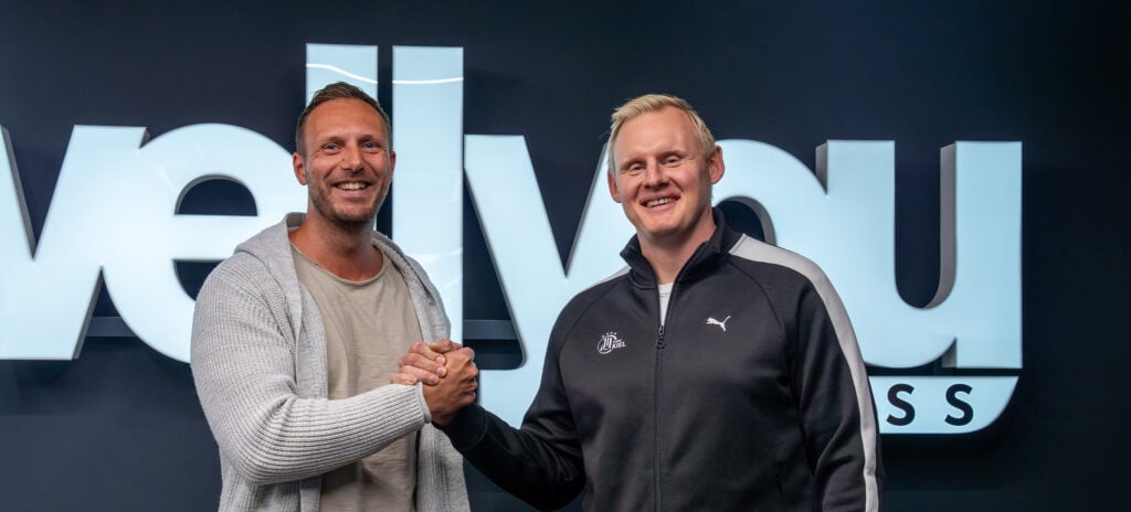 Zwei Männer lächeln und schütteln sich die Hände vor einem großen Schild mit der Aufschrift wellyou. Beide sind leger gekleidet; der eine trägt eine helle Strickjacke und ein Hemd, der andere eine schwarze Puma-Jacke.