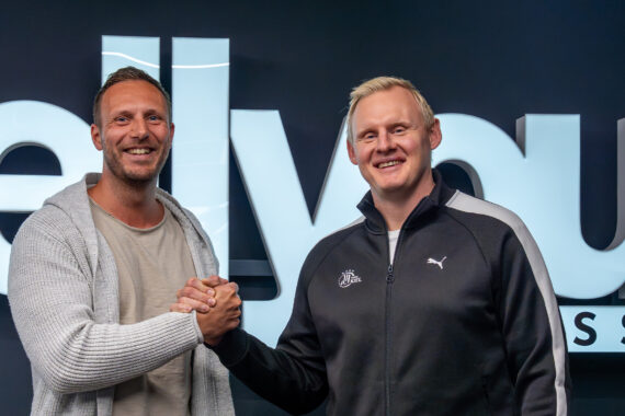 Zwei Männer lächeln und schütteln sich die Hände vor einem großen Schild mit der Aufschrift wellyou. Beide sind leger gekleidet; der eine trägt eine helle Strickjacke und ein Hemd, der andere eine schwarze Puma-Jacke.