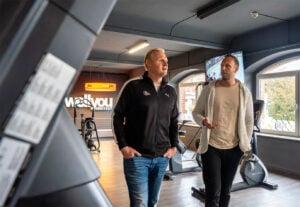 Zwei Männer gehen und unterhalten sich in einem modernen Fitnessstudio mit Fitnessgeräten und großen Fenstern. Das Fitnessstudio ist mit WaiYou Fitness-Schildern und einem Fernsehbildschirm im Hintergrund versehen.
