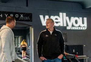 Zwei Männer unterhalten sich in einem Fitnessstudio in der Nähe eines Schildes mit der Aufschrift "Well you FITNESS". Ein Mann trägt eine schwarze Jacke, der andere einen grauen Kapuzenpulli. Im Hintergrund trainiert eine Frau in der Nähe eines HYROX AREA-Schildes.