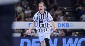 Ein Handballspieler in einem schwarz-weiß gestreiften Trikot jubelt leidenschaftlich auf dem Spielfeld, mit offenem Mund und geballten Fäusten. Im Hintergrund sind Zuschauer zu sehen.