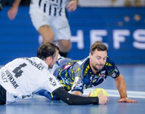 Zwei männliche Handballspieler springen auf das Spielfeld und greifen nach einem gelben Ball; der eine trägt ein weißes Trikot, der andere ein dunkelblau gemustertes Trikot. Der Hintergrund ist unscharf, der Text teilweise sichtbar.