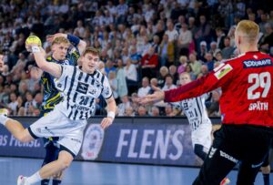 Ein Handballspieler in einem weiß-schwarzen Trikot springt, um den Ball in Richtung Tor zu werfen, während ein Verteidiger in Blau versucht, ihn zu blockieren, und ein Torwart in einem roten Trikot sich darauf vorbereitet, den Schuss zu stoppen. Die Tribünen sind mit Zuschauern gefüllt.