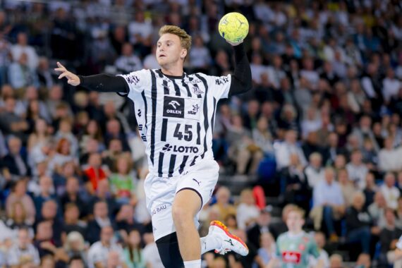 Ein Handballspieler in einem schwarz-weiß gestreiften Trikot mit der Nummer 45 springt in die Luft, um einen gelben Ball zu werfen, während im Hintergrund eine unscharfe Menschenmenge zusieht.