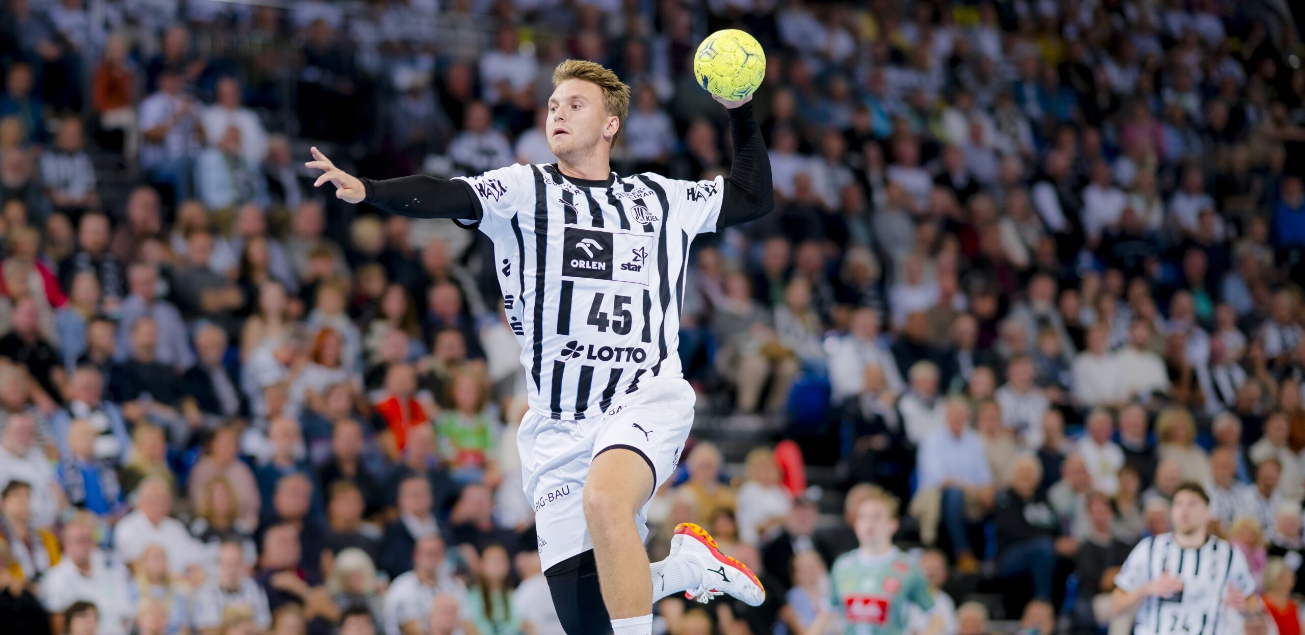 Ein Handballspieler in einem schwarz-weiß gestreiften Trikot mit der Nummer 45 springt in die Luft, um einen gelben Ball zu werfen, während im Hintergrund eine unscharfe Menschenmenge zusieht.