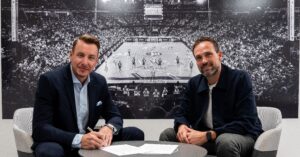 Zwei Männer sitzen an einem Tisch, einer unterschreibt Dokumente, beide lächeln in die Kamera. Hinter ihnen ist ein großes Schwarz-Weiß-Foto einer überfüllten Basketball-Arena zu sehen.