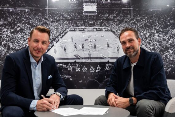 Zwei Männer sitzen an einem Tisch, einer unterschreibt Dokumente, beide lächeln in die Kamera. Hinter ihnen ist ein großes Schwarz-Weiß-Foto einer überfüllten Basketball-Arena zu sehen.