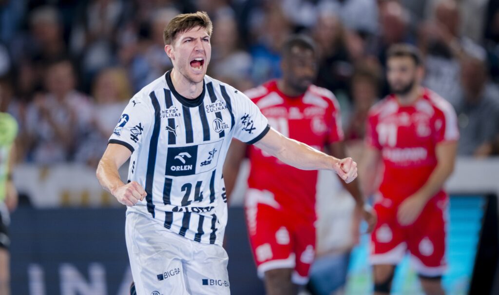 Ein männlicher Handballspieler in einem weiß-blau gestreiften Trikot jubelt mit geballten Fäusten und offenem Mund während eines Spiels. Die gegnerischen Spieler in roten Trikots sind im unscharfen Hintergrund zu sehen.