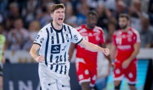 Ein männlicher Handballspieler in einem weiß-blau gestreiften Trikot jubelt mit geballten Fäusten und offenem Mund während eines Spiels. Die gegnerischen Spieler in roten Trikots sind im unscharfen Hintergrund zu sehen.