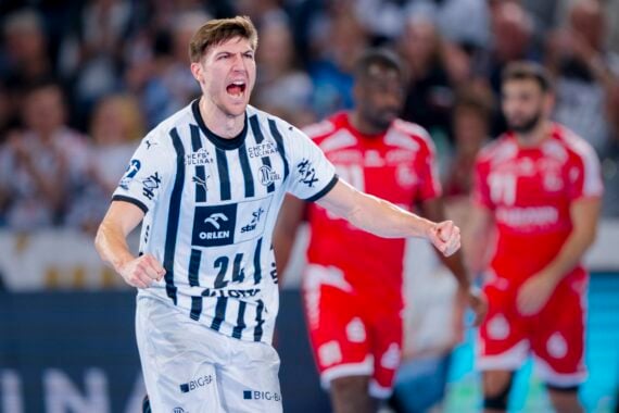 Ein männlicher Handballspieler in einem weiß-blau gestreiften Trikot jubelt mit geballten Fäusten und offenem Mund während eines Spiels. Die gegnerischen Spieler in roten Trikots sind im unscharfen Hintergrund zu sehen.